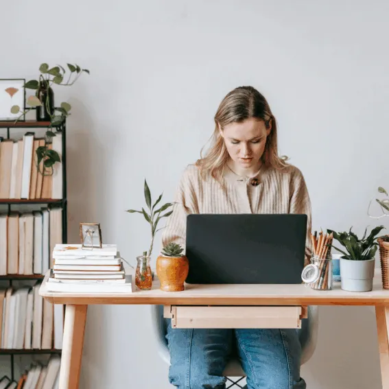 Light office Frau sitzt vor ihrem Laptop an einem Holzschreibtisch. Neben ihr stehen einige Bücher und hinter ihr ein Bücherregal.