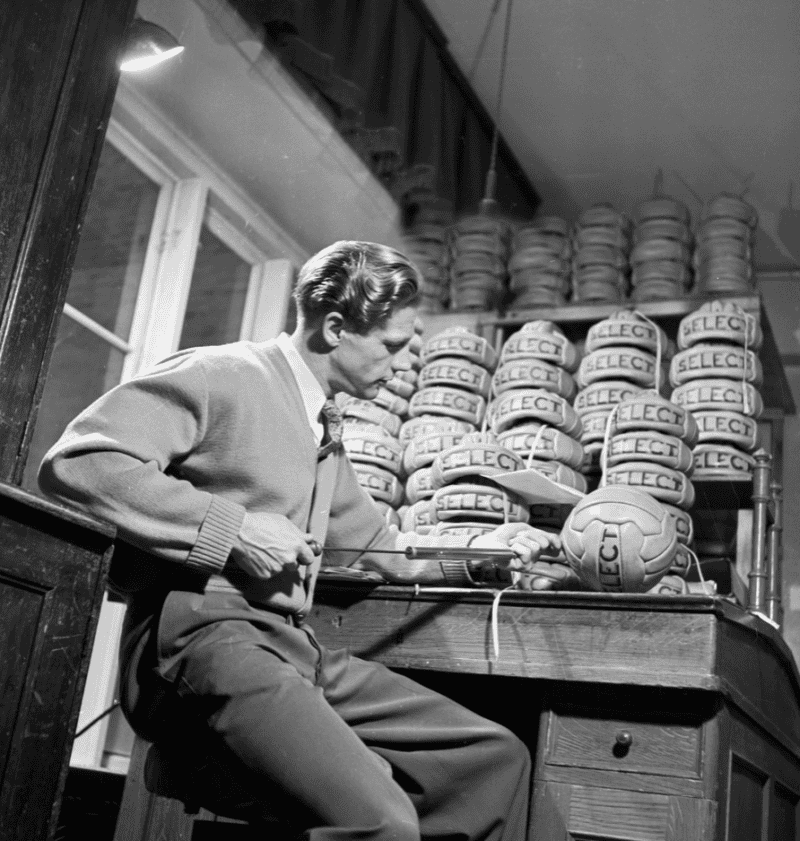 A man is sitting at a desk with Select footballs. He is filling one of them with air with a pump.