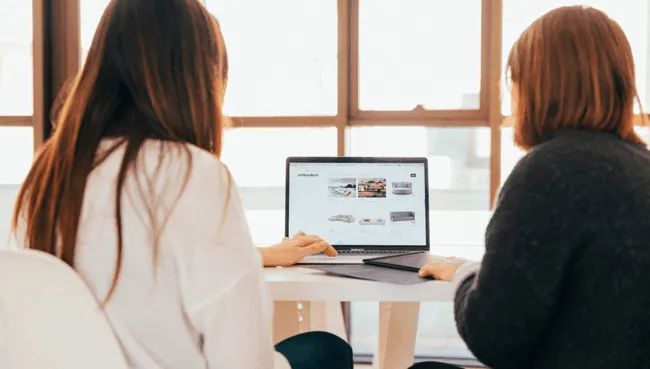 Two women sitting in front of a laptop, backs to the camera.