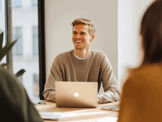 Smilende medarbejder i møde med kolleger, arbejder på bærbar computer — Kontainer Digital Asset Management (DAM) og Product Information Management (PIM) løsninger.