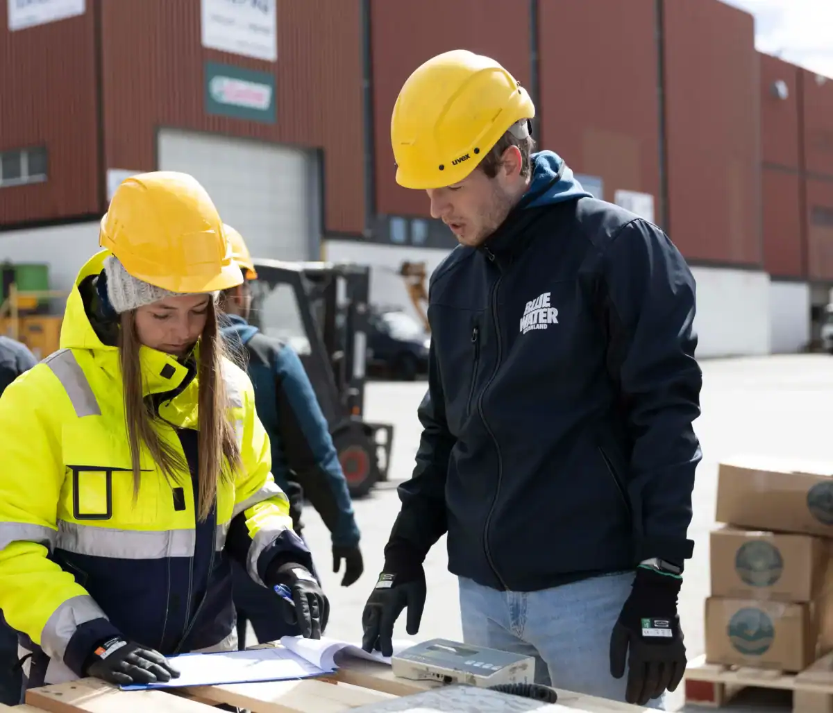 Logistics_BWS_Kontainer-DAM Two Blue Water Shipping employees looking at paperwork atop a pallet outside a storage warehouse