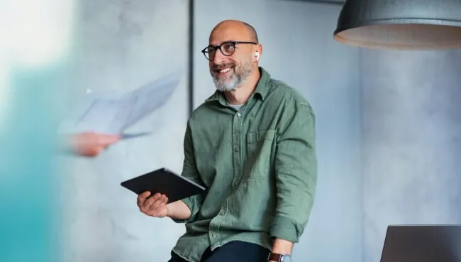 A cheerful man wearing glasses holding a tablet, interacting in a modern office environment. The image portrays professionalism and a collaborative atmosphere. Ideal for business, technology, or coworking-related themes.
