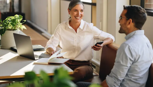 Businesswoman sitting at desk and explaining new project to coworker. Mature woman discussing working with partner in coworking office.